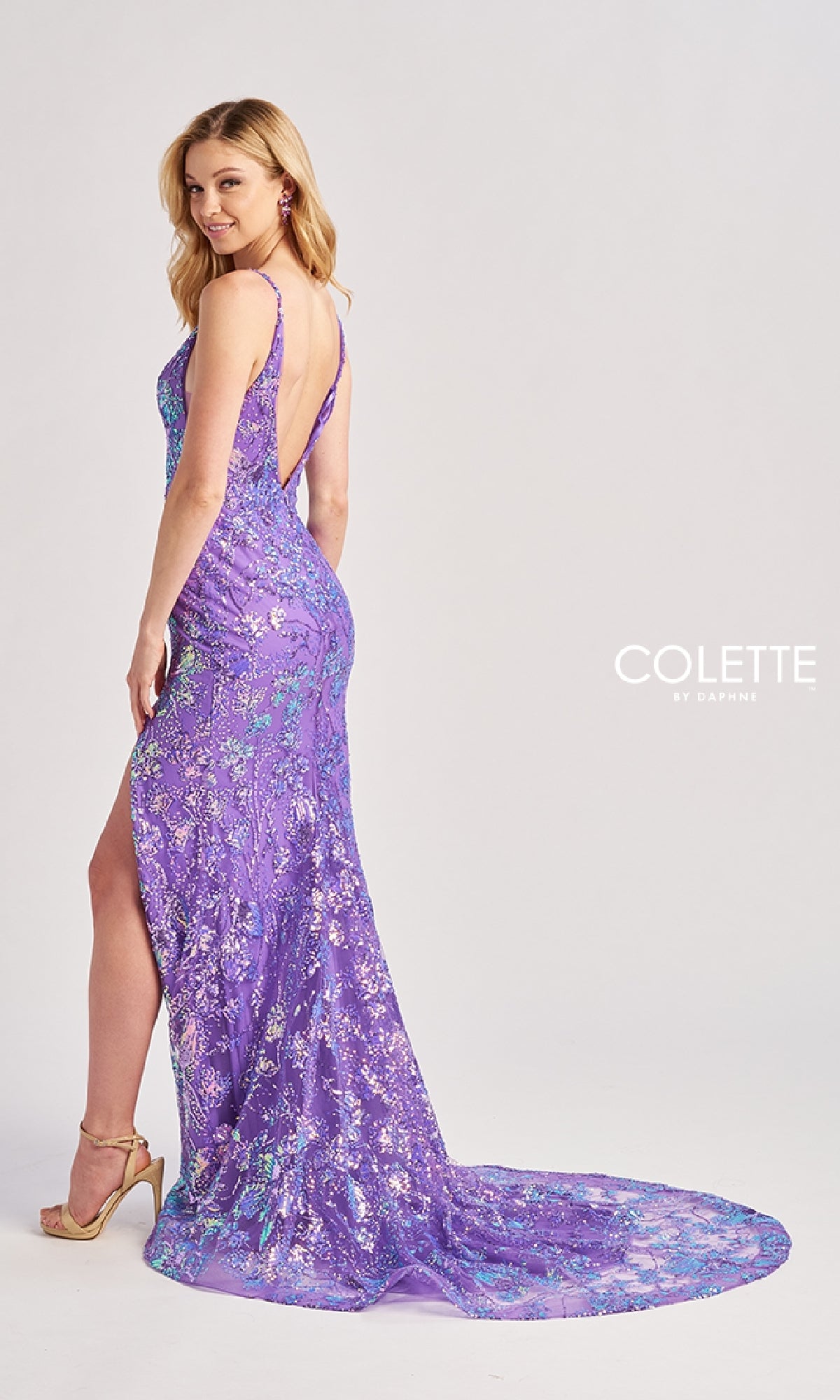 Wearing the Colette CL8430 long prom dress—sleeveless, floor-length, with a thigh-high slit, deep back, train, and floral sequin embroidery—a woman stands in heels looking over her shoulder, radiating elegance.